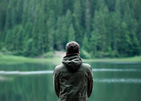 pexels-photo-1172207-1172207 A person in a green jacket stands by a serene forest lake, capturing calming nature.